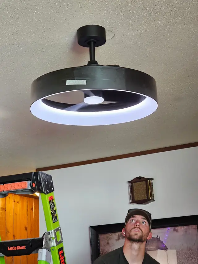 A Thunder Electric electrician on a ladder looking up at a black LED ring ceiling fan he just installed